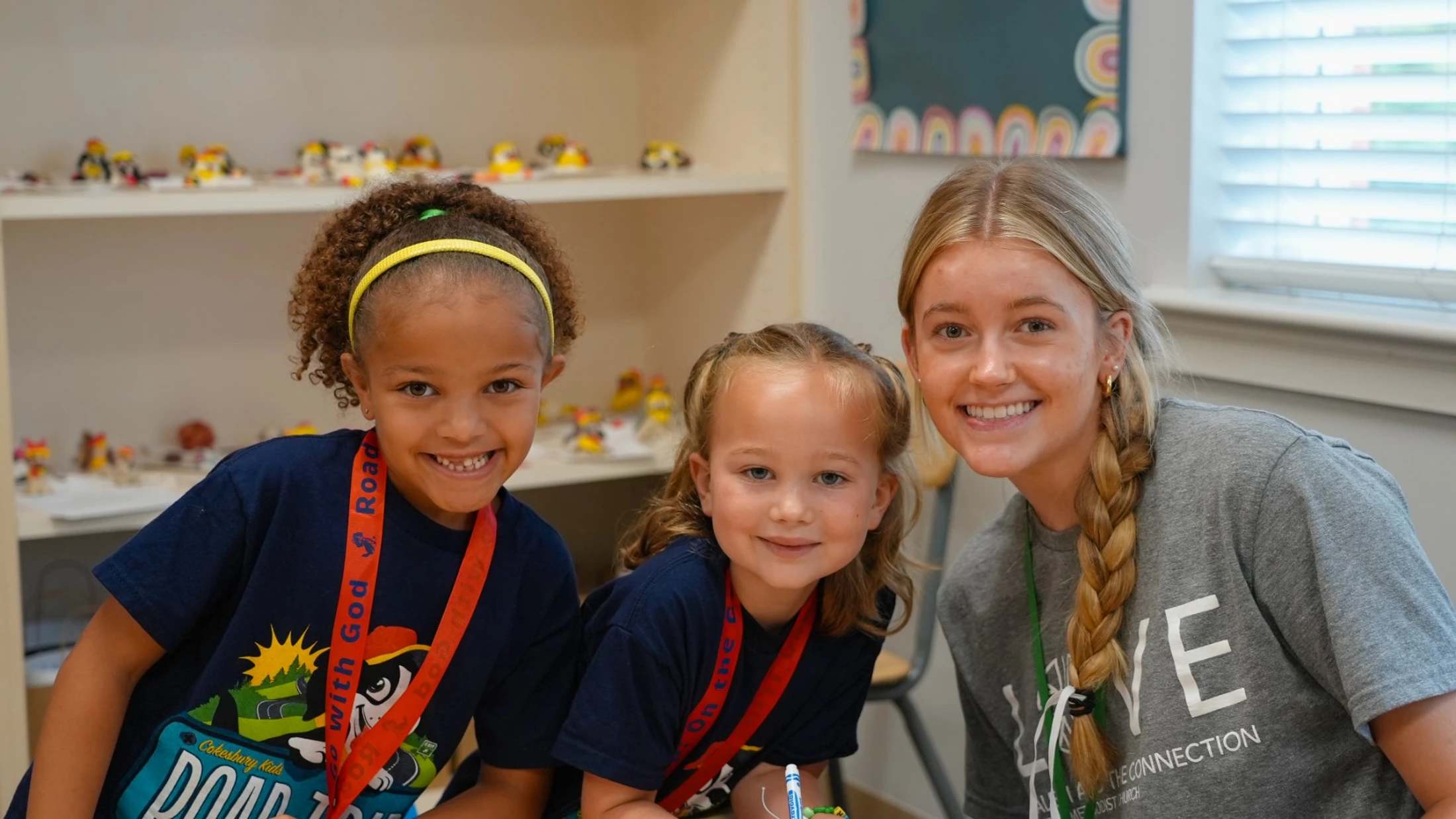 A female camp counselor with a blonde braid smiles with two young girls in a classroom. They are all looking at the camera during a camp activity.
